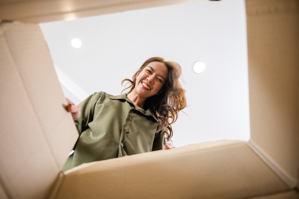 A smiling woman unpacks a cardboard box, her joyful expression suggesting a positive moving experience. She wears a green shirt in a bright, homey setting. Local movers make moving day easier!