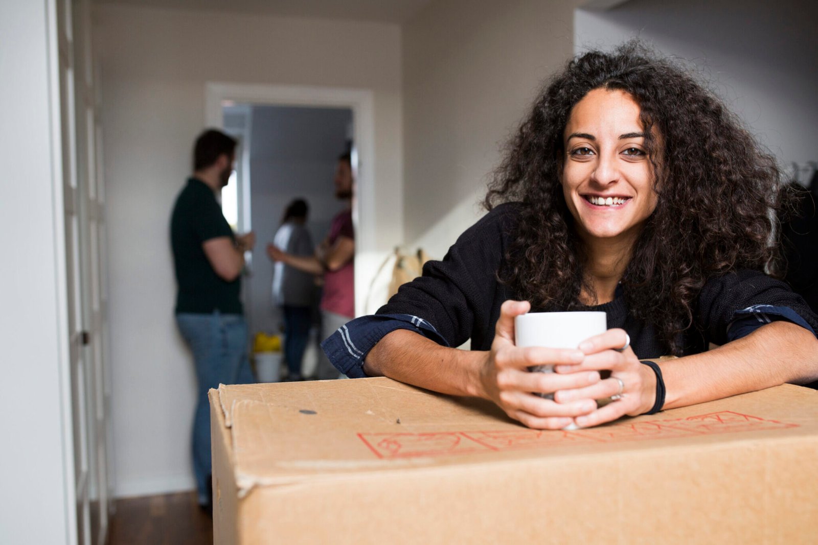 Smiling young woman with curly brown hair sits on a cardboard box, holding a white mug, while local movers unpack in the background. She looks directly at the camera.