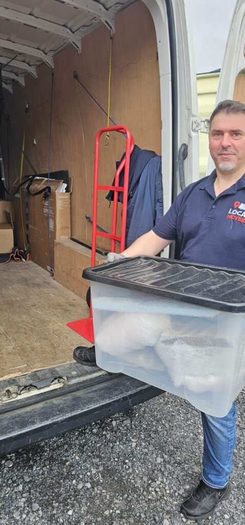 Man in a blue polo shirt with "Local Movers" logo carries a clear plastic bin filled with white items, standing beside a moving van, ready for moving from Dublin to Sligo.