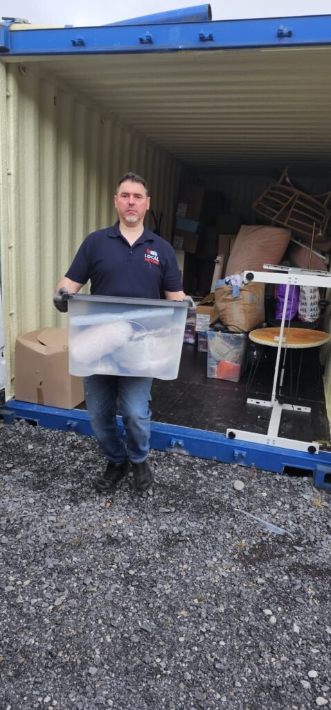 Man in a blue polo shirt and jeans, wearing gloves, holds a large clear plastic bin filled with items while standing in front of an open shipping container packed with boxes and furniture, ready for moving from Dublin to Sligo.