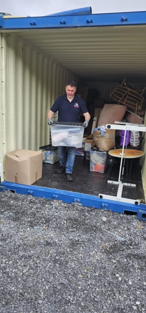 Man loading a clear plastic bin filled with items into a shipping container, possibly part of moving from Dublin to Sligo. The container is packed with furniture and boxes.