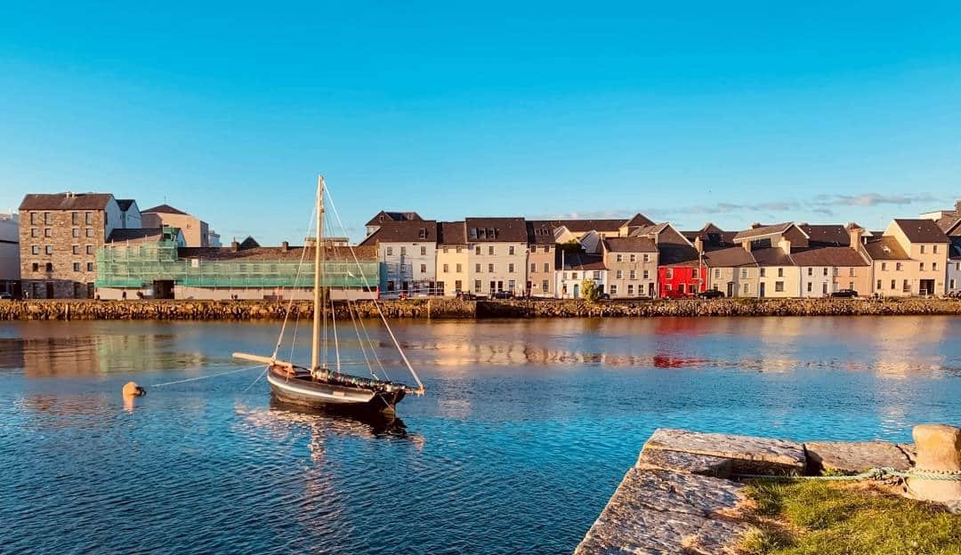 A traditional sailboat floats on the calm, blue waters of Galway harbor, with colorful terraced houses lining the waterfront in the background under a clear sky.