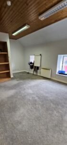 Empty office space with grey carpet, wooden ceiling, whiteboard, and bookshelf, ready for commercial relocation.