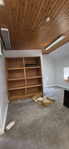 An empty office space with a wooden ceiling, a large wooden bookshelf, and gray carpet. Cardboard boxes and packing materials are scattered on the floor near the bookshelf.