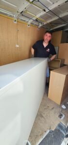 A man in a black polo shirt with the "Local Movers" logo helps move a large white desk inside a moving van, showcasing professional commercial relocation services.