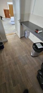 Newly installed wood-look vinyl flooring in a room with an open white door, a gray speckled countertop, and a recycling bin, suggesting a recent office move or renovation.