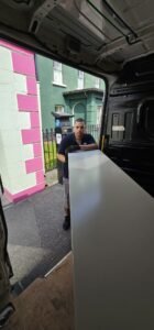 A man wearing gloves holds a large white desk in the back of a moving van parked on a street with a pink and white building and a black fence.