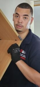 A professional office mover wearing black Velway gloves and a navy blue polo shirt with a "Local Movers" logo carefully carries a light brown wooden furniture piece.