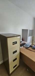 A beige and brown metal filing cabinet stands in an office setting next to a wooden desk with a computer monitor and papers, suggesting a commercial relocation.