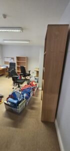 Office relocation in progress: boxes and office supplies are neatly organized on the floor next to a tall, empty wooden bookshelf, with office chairs and desks visible in the background.