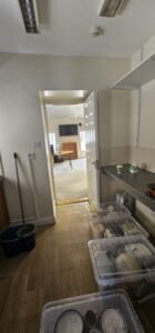 Empty room with hardwood floors, open door leading to a living room with a TV and desk, and clear plastic storage boxes filled with dishes on the kitchen counter.