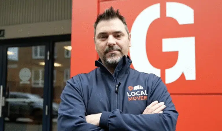 A confident man with a beard and spiky hair stands with his arms crossed in front of a bright red building with a large white 'G'. He wears a navy blue jacket with a "Local Mover" logo, emphasizing our moving licenses, insurance & safety standards.