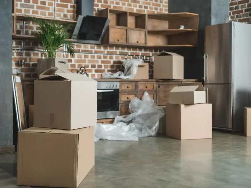 Cardboard boxes stacked in a partially unpacked kitchen, indicating a recent move. The room features exposed brick, wooden cabinets, and stainless steel appliances. Bubble wrap and packing materials are scattered on the floor, suggesting ongoing relocation efforts by local movers.